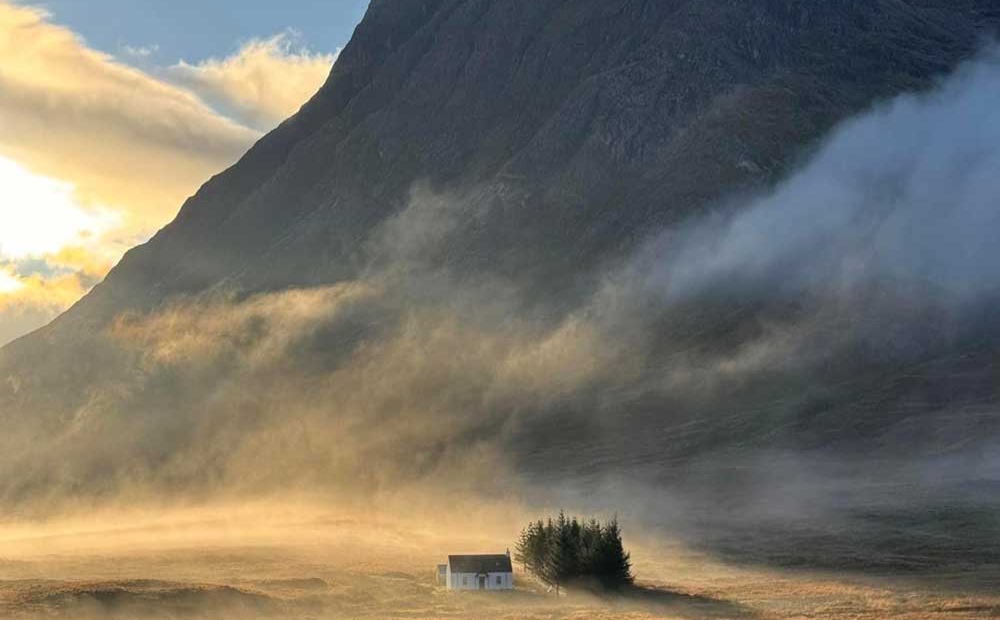 Glen Coe in Scotland with a white cottage below a mountain with mist and sunshine in the background